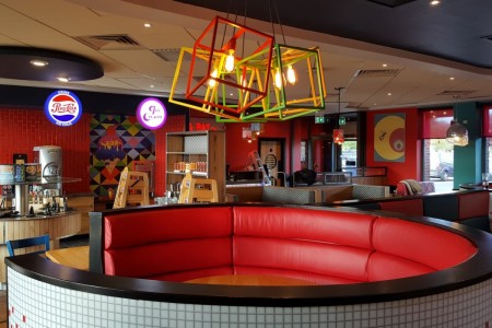 Pizza Hut, Stevenage - round booth with red seating and tiled exterior. Geometric bright lampshades and various neon signs in background 