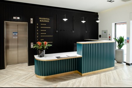 Prudential Buildings, Bristol - light wooden floor, seating area with small tables, teale reception desk, black panelling behind desk with floor numbers on, black vase on desk with red and green flowers
