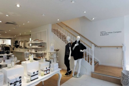 The White Company, Stamford - white table with white candles at foot of stairs, mannequins next to stairs wearing black clothing, wooden staircase going up