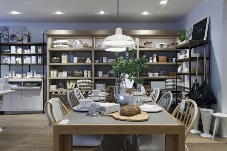 The White Company, Norwich - large wooden table with 6 chairs, on the table there's fake food and plants, and crockery set out. Behind is shelving with a variety of white products