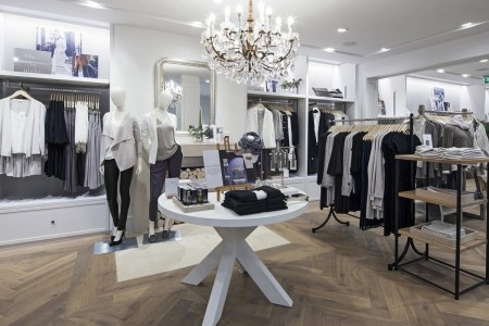 The White Company, Norwich - hardwood parquet floor with white table featuring clothes, clothing rails around the centre table, large chandelier over table
